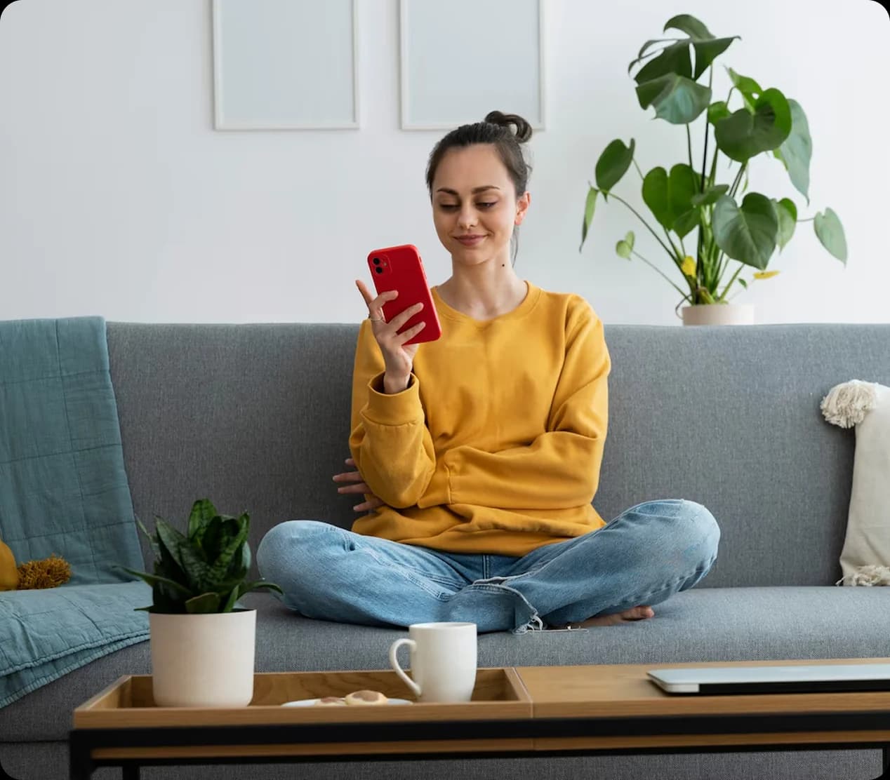 Smiling woman sitting on a sofa using smartphone at home with plants and coffee table.